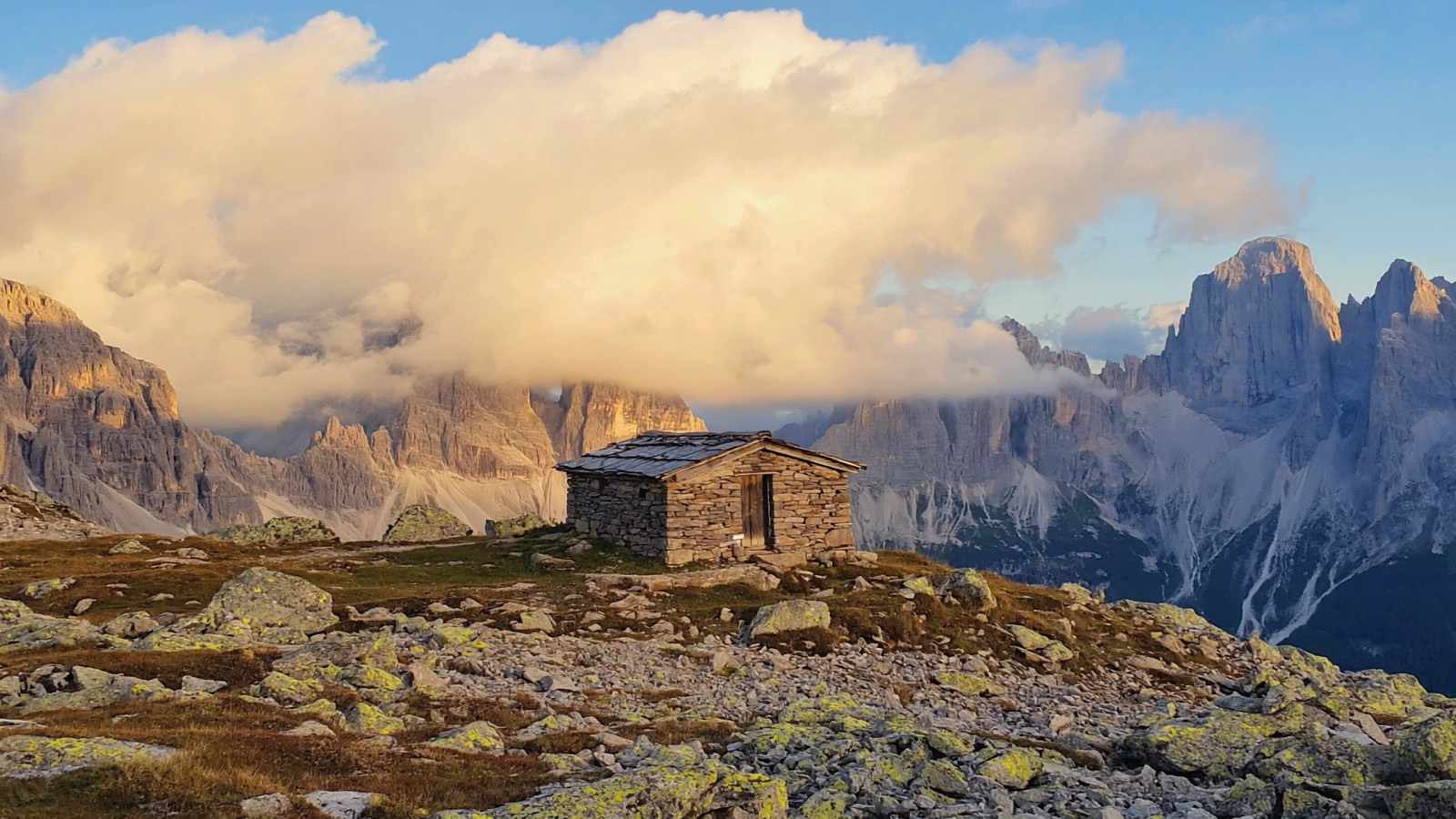 Steinhütte auf einem Grenzsattel zwischen zwei Tälern, dahinter kahle Grasrücken und ferne Felsgipfel im Nachmittagslicht
